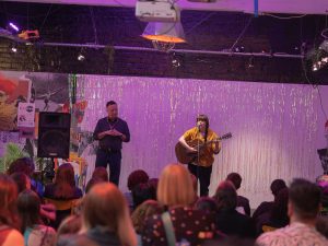 a white queer woman with short brown hair sings on a mic with a guitar in front of a seated audience