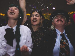 three white people look upwards with smiles and eyes closed as they enjoy glitter being thrown over theirselves in a dark party setting